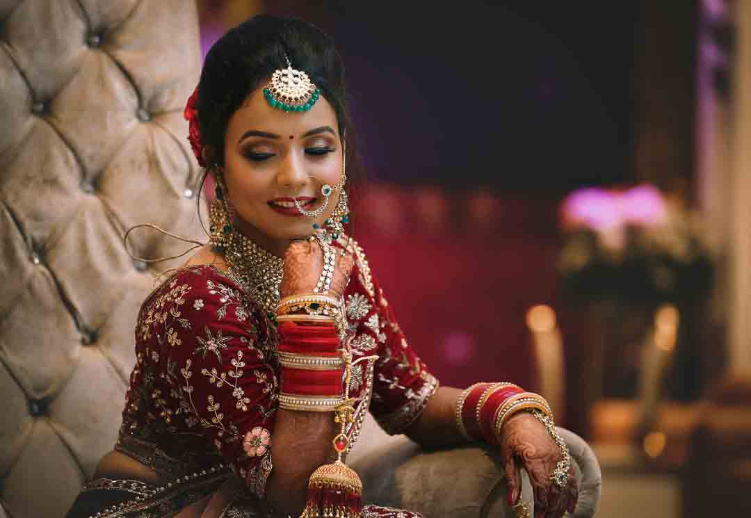 A Bihari bride seated gracefully in a maroon bridal lehenga, adorned with heavy jewelry, traditional red bangles, and an elegant smile, captured in a regal indoor setting.