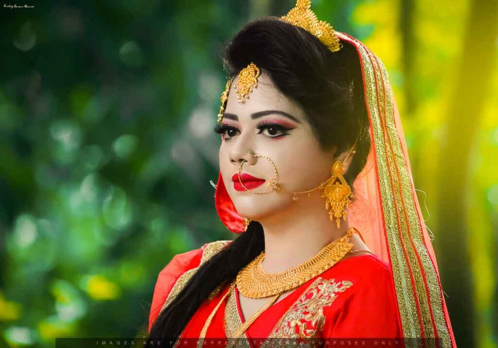 A beautiful Muslim bride in a vibrant red traditional outfit adorned with golden jewelry, including a large nose ring, earrings, and a forehead ornament, captured outdoors with a soft green forest background and glowing sunlight.