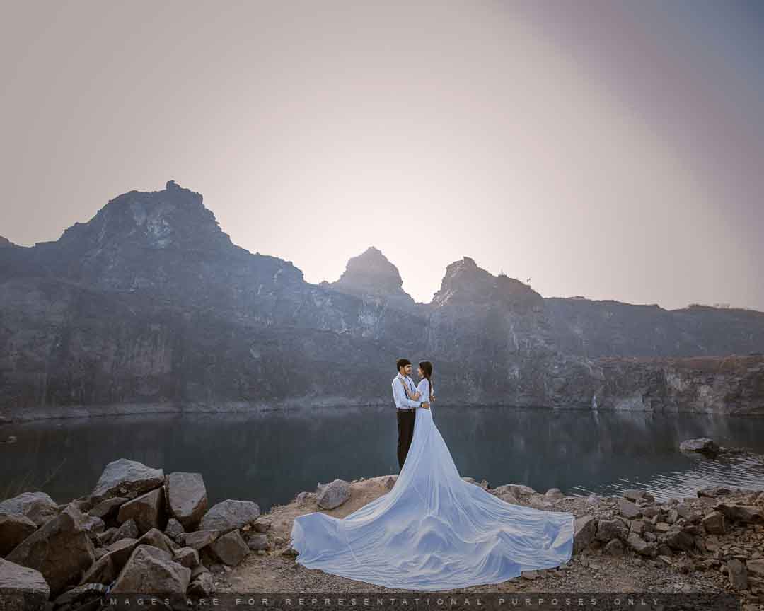 Couple posing romantically on a rocky lakeside with mountains in the background and a long flowing gown trailing behind.
