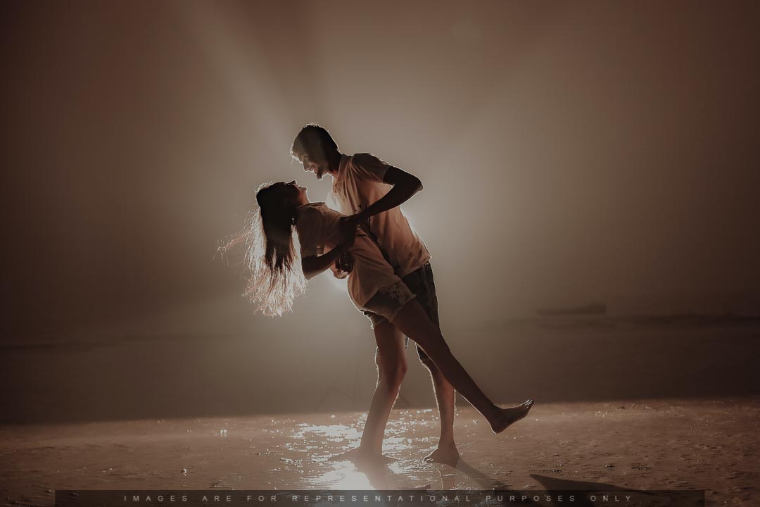Romantic pre-wedding photoshoot of a couple dancing on a beach with dramatic backlighting.