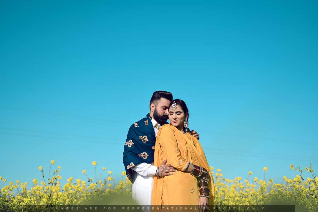 Couple in traditional attire posing in a mustard field under a bright blue sky during a pre-wedding shoot.