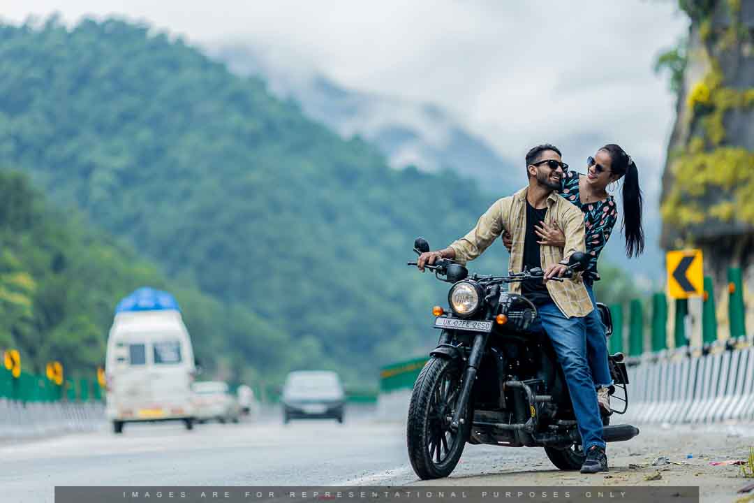 Pre-wedding photo of a fun-loving couple riding a Royal Enfield on a scenic mountain highway.